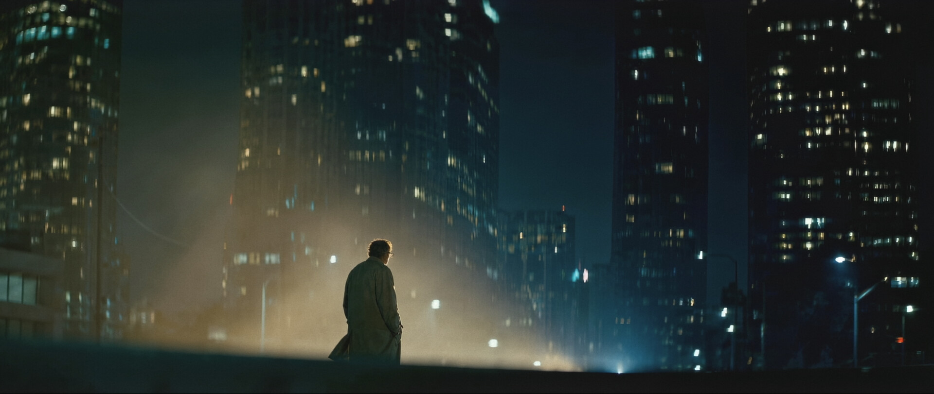 Still from What Is Freedom — a lone man in a trench coat standing on an empty overpass before a wall of lit skyscrapers at night.